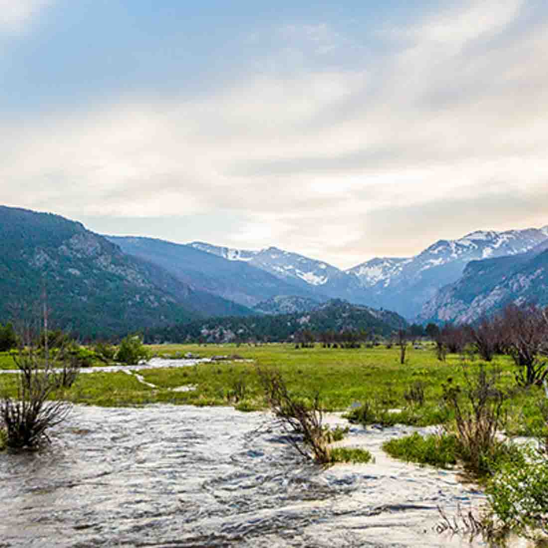 A serene landscape with a stream in the foreground, green grass, and snow-capped mountains in the background under a cloudy blue sky.
