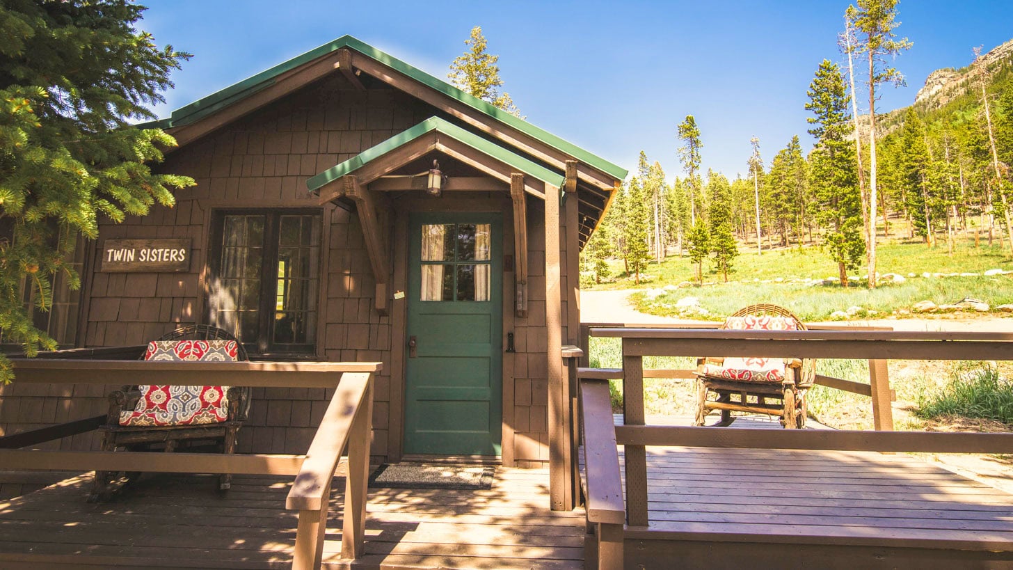 A small wooden cabin with a green door and deck, surrounded by trees and mountains.